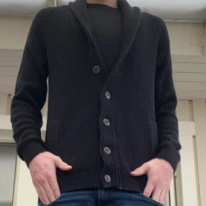 Svart stickad kofta med knappar - En Tvärfet Svart stickad kofta med snygg sjalkrage och stora knappar framtill. Långärmad modell med ribbade muddar och en klassisk, enkel look som passar till jeans eller över en t-shirt.