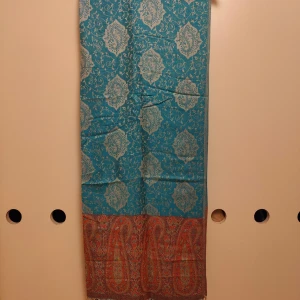 Turkos paisleymönstrad sjal med fransar - Snygg turkos sjal med paisleymönster i beige och detaljerad bård i rött, orange och grönt. Nederkanten har långa, tvinnade fransar som ger extra stil. Perfekt att slänga över axlarna för en färgklick till din outfit.