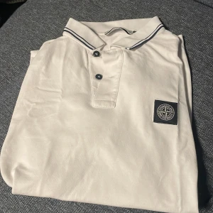 Stone island pike - Vit pikétröja från Stone Island med klassisk krage, svarta knappar och ikonisk logotyp på bröstet. Slim fit-modell med diskreta svarta ränder på kragen. Perfekt för dig som gillar stilrena och exklusiva plagg. Skick 10/10. Pris kan diskuteras. Nypris- 2000. 