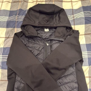 Svart pufferjacka med huva från McKinley - Svart pufferjacka från McKinley i storlek Large. Jackan har quiltad framsida och rygg, huva och hel dragkedja. Materialet är syntet och polyester, vilket gör den lätt och skön. Perfekt för kyliga dagar när du vill ha en stilren look.