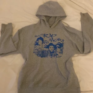 Grå Mount Rap More hoodie från Gildan - Säljer en grå hoodie från Gildan med blått Mount Rap More-tryck på bröstet. Tröjan har huva och en stor magficka framtill. Perfekt för dig som gillar streetwear och vill sticka ut med ett unikt motiv.