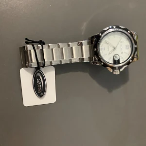 Skone klocka med Datum  - Stilren klocka från Skone med rund urtavla i vitt och markerade siffror. Armbandet är i silverfärgat metall och har en klassisk länkdesign. Klockan har detaljerad boett och tydlig datumvisning. Perfekt för dig som gillar en clean och modern look.