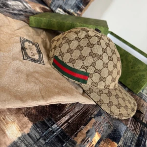 Gucci keps med GG-mönster och läder - Exklusiv Gucci keps i beige och brunt med klassiskt GG-monogram över hela kepsen. Har grönt och rött band på sidan och justerbar rem i brunt läder baktill. Tillverkad i bomull med detaljer i läder, perfekt för dig som gillar lyxig streetstyle.
