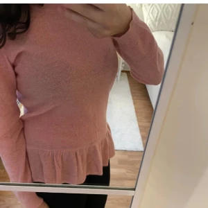 Rosa stickad tröja med volang - Supersöt ljusrosa stickad tröja med volangkant nedtill och långärmad design. Tröjan har en snygg dragkedja i nacken som detalj och sitter normalt på kroppen. Perfekt för dig som gillar mjuka material och vill ha en feminin touch på din outfit.