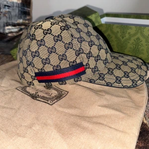 Gucci keps med GG-mönster och röd-blå detalj - Snygg beige keps från Gucci med klassiskt GG-monogram i mörkblått över hela kepsen. På sidan finns en ikonisk röd och marinblå rand. Justerbar rem baktill och böjd skärm. Perfekt för dig som vill ha en lyxig och trendig accessoar.