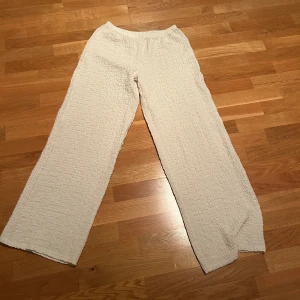 Beiga ribbade byxor från Gina Tricot - Säljer ett par beiga byxor från Gina Tricot i storlek M. De har en ribbad, texturerad yta och är tillverkade i ett mjukt syntetmaterial. Byborna har elastisk midja och ett loose, wide fit som ger en chill vibe. Lowwaist