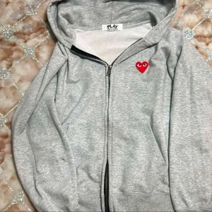 Grå hoodie Comme des Garçons Play - Grå hoodie från Comme des Garçons Play med dragkedja och huva. Ikoniskt rött hjärta med ögon broderat på bröstet. Mjuk bomullskänsla och klassisk passform, perfekt för en chill och stilren look.