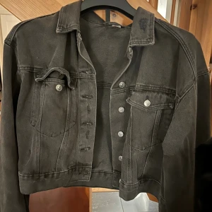 Svart croppad jeansjacka från Divided - Säljer en svart croppad jeansjacka från Divided med klassisk krage, silverfärgade knappar och två bröstfickor. Jackan har en loose passform och är tillverkad i slitstarkt jeanstyg. Perfekt för dig som gillar streetstyle och vill ha en cool look.