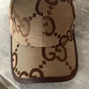 Gucci beige keps med GG-mönster - Snygg keps från Gucci i beige och bruna toner med det klassiska GG-logomönstret över hela kepsen. Kepsen har böjd skärm med brunt kantband och är tillverkad i slitstark bomull. Perfekt för dig som vill ha en lyxig och trendig accessoar.