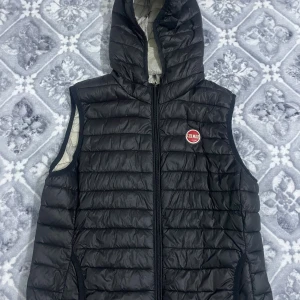 Svart dunväst från Colmar med huva - Svart dunväst från Colmar med quiltad design och huva. Västen har en dragkedja framtill och ett Colmar-märke på bröstet. Insidan är ljus och västen har en clean, sportig look som funkar till många stilar.