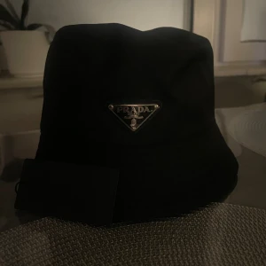 Prada bucket hat - Stilren svart bucket hat från Prada med ikonisk metalltriangel framtill. Hatten är tillverkad i blankt syntetmaterial som ger en lyxig känsla. Perfekt accessoar för dig som vill ha en clean och trendig look. Märkesetikett och tagg medföljer.