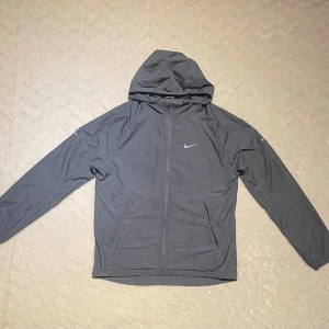 Nike Miller grå - Snygg grå vindjacka från Nike Running med huva och dragkedja framtill. Jackan har två sidofickor och ett stilrent Nike Swoosh-tryck på bröstet. Perfekt för dig som gillar sportig stil och vill ha något lätt och funktionellt.