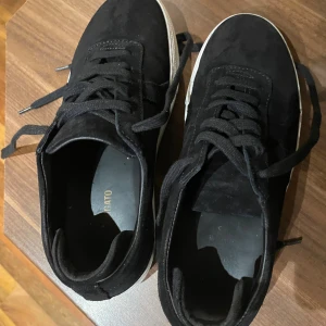Svarta sneakers från ARIGATO i mocka - Snygga svarta sneakers från ARIGATO med ovandel i mjuk mocka och klassisk vit platt sula. Skorna har rund tå, svarta snören och diskret guldfärgad logga på sidan. Perfekt för dig som gillar stilrena och trendiga sneakers med lite extra premiumkänsla.