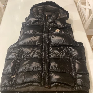 Svart dunväst från Moncler med huva - Svart, glansig dunväst från Moncler med huva och två fickor med dragkedja. Klassisk Moncler-logga på bröstet och coolt serietryck på insidan. Västen har quiltad design och hel dragkedja framtill. Perfekt för att maxa din streetstyle. Nfc tagg funkar, du kan lista ut varför den är billig, den har även en brun liten fläck på insidan men man ser ju inte den utefrån 