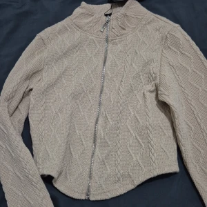 Beige stickad kofta från Amisu - Beige stickad kofta från Amisu med snyggt kabelstickat mönster och hel dragkedja framtill. Koftan har hög krage och långa ärmar, perfekt för lager-på-lager. Mjuk och skön känsla, passar till allt.