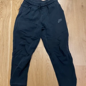 Svarta Nike tech mjukisbyxor  - Svarta mjukisbyxor från Nike med dragsko i midjan och diskret Nike-logga på låret. Byxorna har sidofickor och är tillverkade i mjukt material som är perfekt för chill eller träning. Passformen är normal och benen är något avsmalnande nedtill.