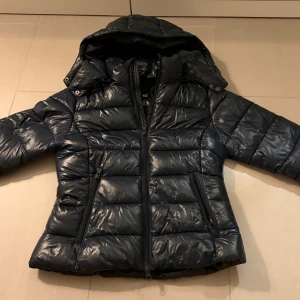 Mörkblå pufferjacka med huva - Säljer en mörkblå, glansig pufferjacka från lager 157. Jackan har avtagbar huva, dragkedja framtill och två fickor med dragkedja. Den är vadderad och har en klassisk, lite kortare passform som är perfekt för kyliga dagar.