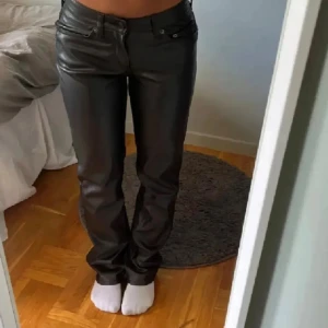 Svarta raka byxor i skinnimitation - Snygga svarta byxor i skinnimitation med klassisk femficksdesign och raka ben. Byxorna har låg midja och en stilren look som passar perfekt till en trendig outfit. Materialet ger en cool känsla och de sitter riktigt bra på kroppen.