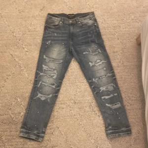 Supply & Demand blå ripped jeans 30W - Blå jeans från Supply & Demand med slitna detaljer och trasiga partier framtill, samt inslag av bandanamönster under vissa hål. Jeansen har normal passform, klassisk femficksdesign och stänkmönster över hela tyget. Perfekt för dig som gillar streetstyle.