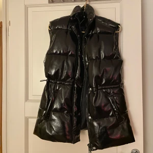 Svart glansig dunväst - Svart dunväst från H&M med glansig finish och hög krage. Västen har justerbar resår i midjan, dragkedja framtill och praktiska fickor. Perfekt för lager-på-lager och ger en cool vibe till din outfit. Jag har haft den hemma i två år ungefär utan att använda den, har endast provat den på. Nypris 449 🖤