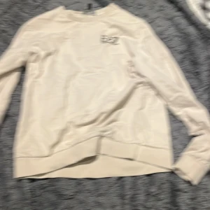 Beige sweatshirt från Emporio Armani - Snygg beige sweatshirt från Emporio Armani med EA7-logga tryckt på bröstet. Tröjan har rund hals, långa ärmar och ribbade muddar vid ärmslut och nederkant. Perfekt för dig som gillar stilrena och klassiska plagg med exklusiv känsla.