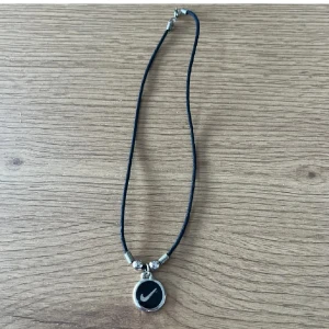 Nike halsband med svart snöre <3 - Stilrent halsband från Nike med svart snöre och silverfärgade detaljer. Hängsmycket är runt med Nike-logga på ena sidan och texten 'NIKE' 