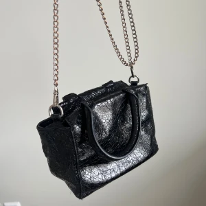 Väska <3 - Liten svart väska med glansig, krackelerad yta. Väskan har både handtag och en lång metallkedja som axelrem, samt dragkedja upptill. Mettalkedjan är något missfärgad på ställen. Från Nelly 🖤