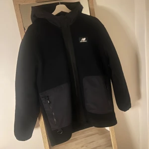 Fleecejacka New Balance - Svart fleecejacka från New Balance med huva och två stora fickor framtill med dragkedja. Jackan har en stilren look med logotyp på bröstet och är tillverkad i mjuk fleece. Perfekt för dig som gillar streetwear och vill ha något varmt och snyggt. Nypris 1650:-