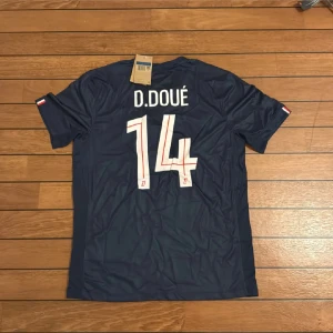 PSG D.Doué matchtröja Nike M - Säljer en officiell Paris Saint-Germain fotbollströja från Nike med D.Doué och nummer 14 på ryggen. Tröjan är mörkblå med rött och vitt geometriskt mönster på framsidan, PSG-emblem och Qatar Airways tryck. Tillverkad i lätt Dri-FIT material för bästa komfort.