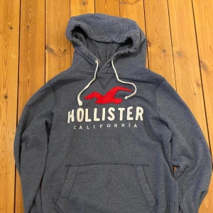 Holister Hoodie  - En hoodie med perfekt passform och premiumkänsla. Inspirerad av klassisk Hollister-stil – avslappnad men stilren. Tillverkad i mjuk bomullsblandning med borstad insida för extra komfort. Ribbstickade muddar och justerbar huva med snörning ger en lyxig street-look. Passform: Regular-fit (sitter snyggt men inte för tight). Storlek: M Design: Enkel, ren logga på bröstet eller ton-i-ton broderi.