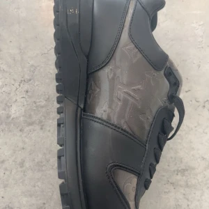 Louis Vuitton svarta sneakers - Snygga svarta sneakers från Louis Vuitton med ikoniskt monogrammönster på sidan. Skorna har robust sula, snörning och är tillverkade i en kombination av skinn och textil. Perfekta för dig som vill ha en exklusiv och stilren look med lyxig känsla.