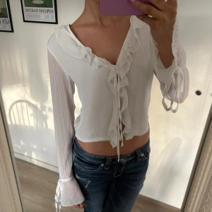 Vit blus med volang och knyt - Säljer en superfin vit blus med långa ärmar med knyt vid ärmslut. Blusen har volangdetaljer längs v-ringningen och framtill, samt knytning i mitten. 💗