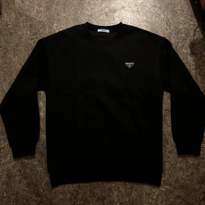 Svart sweatshirt från Prada - Svart crewneck sweatshirt från Prada med ikonisk logotyp på bröstet. Tröjan har ribbade muddar vid hals, ärmslut och nederkant. Tillverkad i mjuk bomull som känns skön mot huden. Perfekt för dig som gillar stilrena designer och exklusiva märken.