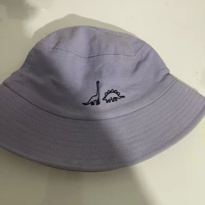 Ljuslila bucket hat med dinosaurier - Säljer en ljuslila bucket hat med broderade dinosaurier framtill. Hatten har klassisk form med bred brätte och är tillverkad i bomull. Perfekt accessoar för att sticka ut med en lekfull touch.