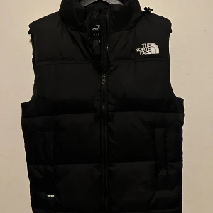 Svart dunväst från The North Face - Svart dunväst från The North Face med hög krage och vit logga på bröstet och ryggen. Västen har en quiltad design, dragkedja framtill och två sidofickor. Perfekt för lager-på-lager och chill stil under kyliga dagar.