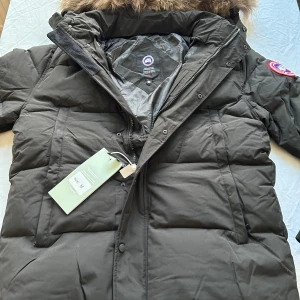 Svart  Canada Goose jacka  - Säljer en svart Canada Goose jacka  med pälskantad huva och klassisk logga på ärmen. Jackan har quiltad design, dragkedja och knappar framtill. Materialet är polyester och bomull, fodrad med dun för extra värme. Perfekt för kalla vinterdagar.