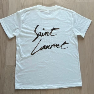 Vit Saint Laurent t-shirt - Helt ny Vit t-shirt från Saint Laurent med svart handskriven logga på ryggen. Klassisk rund hals och korta ärmar. Tillverkad i mjuk bomull för en skön känsla och enkel stil.