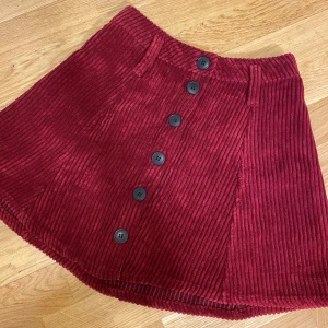 Vinröd manchesterkjol från Bershka - Vinröd kortkjol i manchester från Bershka med markerade breda ränder och snygga svarta knappar framtill. Kjolen har bälteshällor och en lätt A-linjeformad siluett som ger en cool retrovibe.