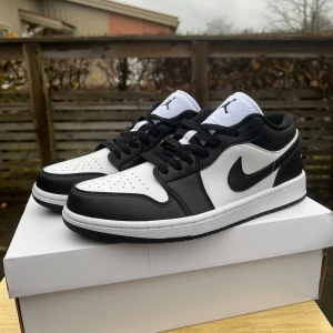 Nike Air Jordan 1 Low svart/vit - Nike Air Jordan 1 Low i svart och vitt skinn med klassisk Swoosh på sidan och Air Jordan-logga på hälen. Lågt skaft, svart snörning och perforerad tåbox för extra stil. Perfekta sneakers för dig som vill ha en clean och tidlös look.