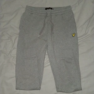 Grå mjukisbyxor från Lyle & Scott - Grå mjukisbyxor från Lyle & Scott med klassisk gul logga på vänster ben. Byxorna har snörning i midjan, två sidofickor och en bakficka. Tillverkade i mjukt bomullsmaterial för maximal komfort. Perfekta för chill eller träning.
