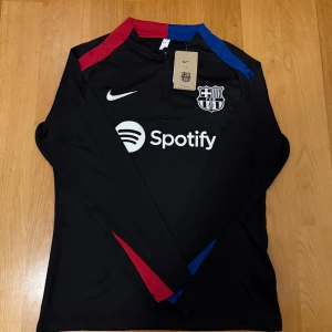 Svart långärmad FC Barcelona tröja - Snygg svart långärmad fotbollströja från Nike med FC Barcelona-logga och Spotify-tryck på bröstet. Tröjan har dragkedja vid halsen och detaljer i rött och blått på kragen och ärmarna. Perfekt för dig som älskar Barca och vill ha en stilren look.