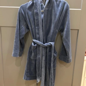 Blå morgonrock med huva från Lindex - Mjuk och chill blå morgonrock från Lindex i storlek XS, har huva, två stora fickor och ett bälte i midjan. Perfekt att slänga på sig efter duschen eller när du bara vill mysa hemma.