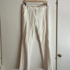 Vita kostymbyxor med raka ben - Snygga vita kostymbyxor med raka ben och diskreta fickor bak. Byxorna har bälteshällor och klassisk passform, perfekta för en clean och stilren look. Materialet känns mjukt och har en lätt glans, vilket ger en fräsch vibe.