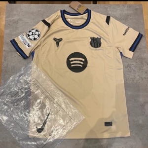 Barcelona Lamine Yamal bortatröja Nike S - Exklusiv FC Barcelona bortatröja med Lamine Yamal och nummer 10. Beige med svarta och blå detaljer, officiella klubbmärken, Champions League-patch och Spotify-logga. Tillverkad i lätt Dri-FIT-material som andas. Perfekt för dig som älskar fotboll och Barca.