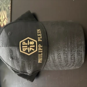 Svart keps från Philipp Plein - Svart keps från Philipp Plein med guldfärgat logotryck och text framtill. Kepsen har en snygg, diskret mönstrad yta med märkets namn ton-i-ton över hela materialet. Justerbar rem baktill med metallspänne. Tillverkad i 100% bomull.