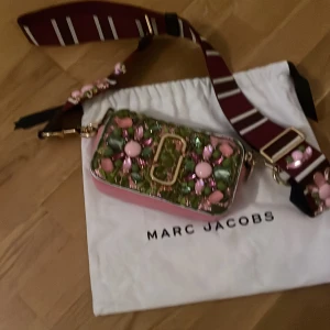 Blommig axelväska från Marc Jacobs - Superfin axelväska från Marc Jacobs med blommigt mönster i grönt och rosa, dekorerad med pärlor och stenar. Väskan har bred, randig axelrem i vinrött och vitt samt detaljer i guld. Tillverkad i skinn med dragkedja och rosa baksida. Helt ny, aldrig använd mått h 11 cm  b18cm bred åt sidan 6