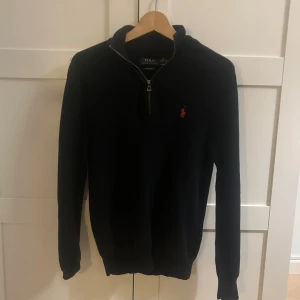 Svart stickad half zip tröja Polo Ralph Lauren - Svart långärmad tröja från Polo Ralph Lauren i mjuk pima bomull. Klassisk half zip-dragkedja och röd broderad logga på bröstet. Perfekt för dig som gillar stilren och tidlös design med en sportig touch.
