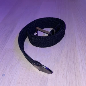 Svart flätat bälte med skinn och metallspänne - Svart flätat bälte med detaljer i skinn och ett stilrent metallspänne. Bältet har en klassisk design med snyggt mönstrad struktur och passar perfekt till jeans eller chinos. Längden är 95 cm och materialet är en kombination av syntet och skinn.