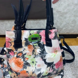 Blommig axelväska med svarta detaljer - Snygg axelväska med blommigt mönster i rosa, orange, grön och svart på vit botten. Väskan har svarta handtag och detaljer i skinnimitation, samt dragkedja och metallspänne framtill. Perfekt för dig som vill sticka ut med färg och mönster.