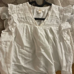 Vit blus med volang från Gina Tricot - Supersöt vit blus från Gina Tricot i storlek 134/140. Blusen har volangdetaljer på axlarna, v-ringad hals och fina broderade partier upptill. Tillverkad i lätt och luftig bomull, perfekt för vår och sommar. Snygg att styla med jeans eller kjol!